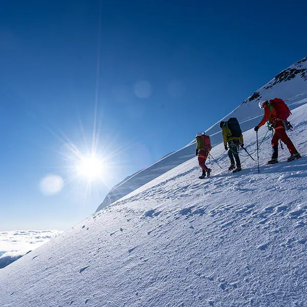 Drie bergbeklimmers die een steile, zonovergoten besneeuwde helling beklimmen.